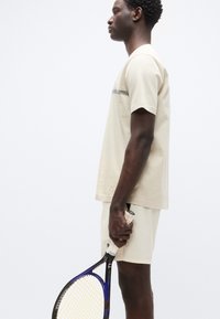 Μπεζ αθλητικό μπλουζάκι με κοντά μανίκια και οριζόντια ρίγα, συνδυασμένο με ταιριαστό σορτς. Το άτομο κρατάει μια ρακέτα του τένις.