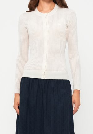 Frau trägt eine enganliegende, weiß gerippte, knöpfbare Strickjacke mit langen Ärmeln und einen dunklen Faltenrock, steht vor einem schlichten Hintergrund.