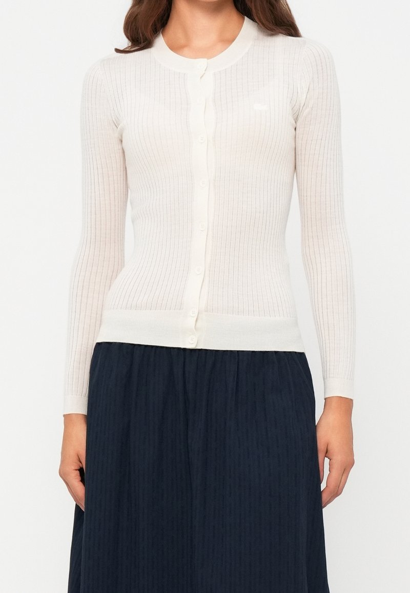 Femme portant un cardigan blanc côtelé ajusté boutonné à manches longues et une jupe plissée foncée, debout devant un fond uni.