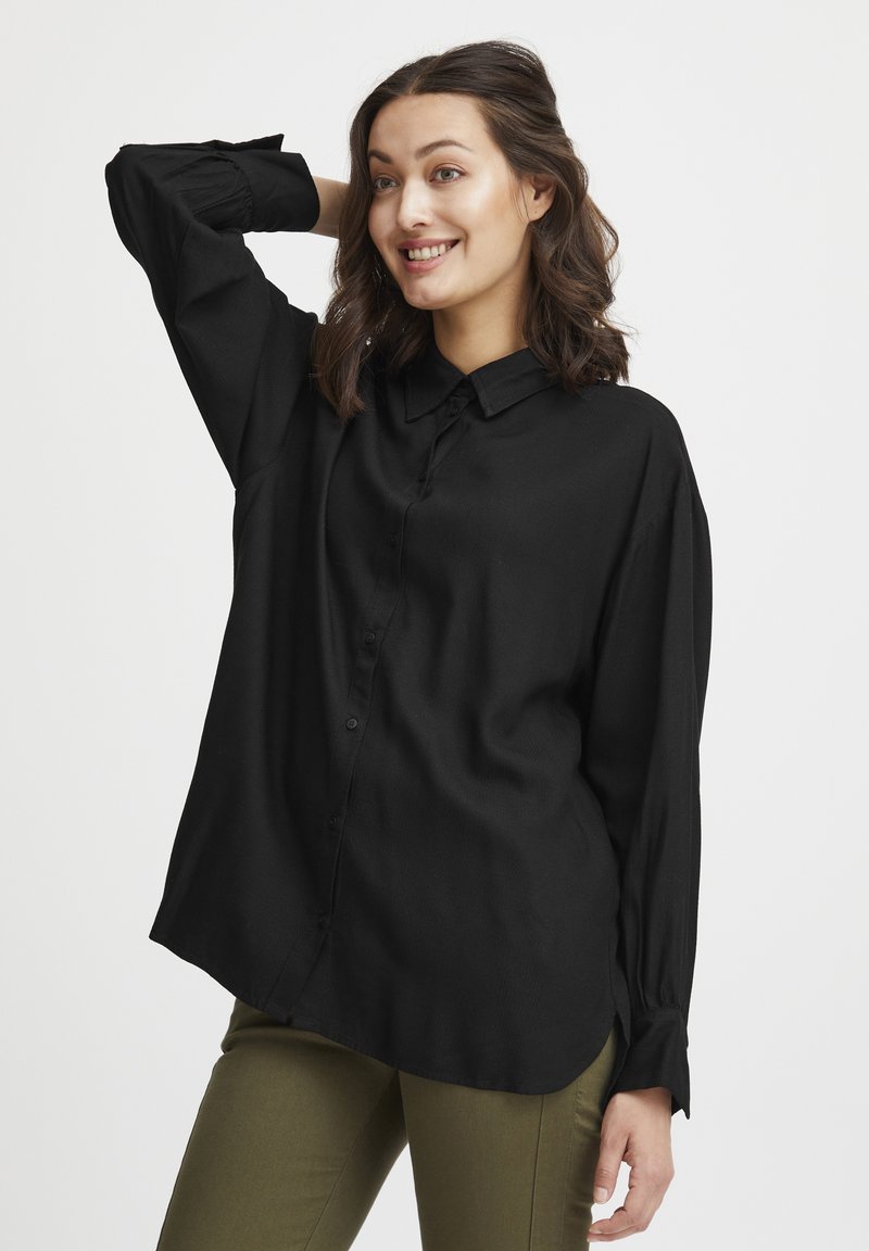 Camicia nera con colletto, maniche lunghe e vestibilità rilassata. Il tessuto sembra liscio con una leggera lucentezza. Indossata con pantaloni verde oliva.