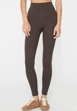 Mujer que lleva leggings marrón oscuro de talle alto y zapatillas de ante marrones con cordones blancos sobre un fondo liso y claro.