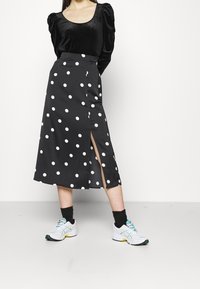 Black midi skirt with white polka dots, featuring a front slit. Paired with a black velvet top and white sneakers with teal accents.