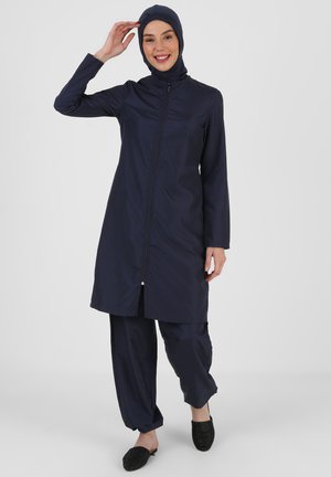 Donna con indosso un completo da bagno modesto blu navy con hijab, top lungo con zip, pantaloni larghi e scarpe slip-on nere, sorridente e che si tocca la testa.