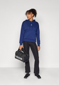 Sweatshirt azul marinho com bolso frontal tipo canguru e pequeno logótipo branco, combinado com calças pretas justas e sapatilhas de desporto pretas. Mala de duffel preta da Adidas.