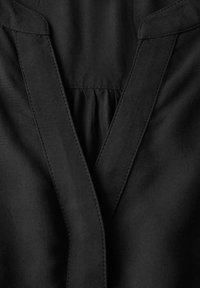 Černá tkanina s výstřihem do V, s nařaseným záhybem uprostřed. Šití je viditelné podél výstřihu a švů.