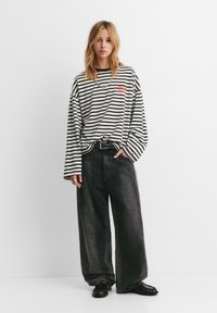 Jonge vrouw die een oversized zwart-wit gestreept shirt met lange mouwen draagt, losse zwarte jeans en donkere schoenen, staand tegen een witte achtergrond.