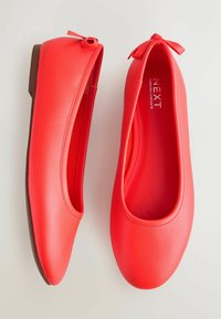 Raudonos plokščios basutės iš lygios dirbtinės odos, suapvalintu priekiu ir lankeliu prie kulno. Pado spalva ruda kontrastui.