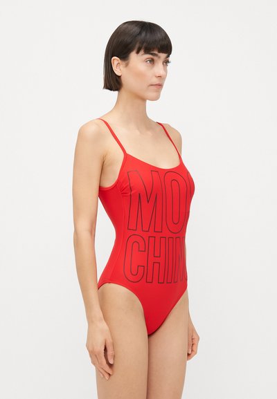 Ženska v rdeči enodelni kopalni obleki z tankimi naramnicami in velikimi črnimi črkami, ki delno berejo "MOSCHINO" spredaj.
