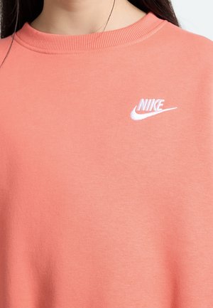 Nærbillede af en person iført en koralpink Nike sweatshirt med et hvidt Nike-logo på venstre øverste bryst.
