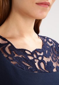 Robe bleu marine avec un col en dentelle florale superposé et un corps en tissu uni texturé. Le modèle met en valeur des motifs délicats en dentelle.