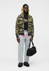 Πράσινο μπουφάν bomber με καμουφλάζ και κίτρινη κουκούλα, μαύρο μπλουζάκι με γραφικό σχέδιο, ανοιχτό μπλε τζιν και μαύρη τσάντα ώμου. Μαύρα παπούτσια με σχέδιο.