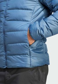 Chaqueta de plumón azul con secciones acolchadas horizontales y bolsillos laterales con cremallera, hecha de un tejido suave y brillante. Pantalones oscuros visibles por debajo.
