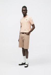 Pikéskjorte i lyserosa med kontraststriper på kragen, kombinert med beige shorts. Tilbehør inkluderer brun belte og svarte sandaler med hvite sokker.