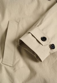 Beige kappaärmsdetalj med knappstängning, kontrasterande svarta knappar och en distinkt flikdesign, tillverkad av ett texturerat tyg.