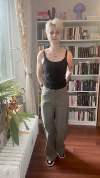 Uma pessoa com cabelo loiro curto, vestindo uma camisola preta sem mangas e calças cinzentas, está num quarto com uma estante e plantas junto a uma janela.