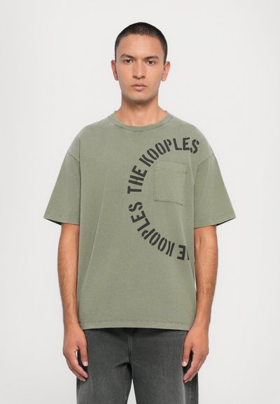 Ελαιόχρωμο oversized t-shirt με μπροστινή τσέπη, με μαύρο κυκλικό γραφικό κείμενο "THE KOOPLES" στην αριστερή πλευρά. Κατασκευασμένο από βαμβάκι.