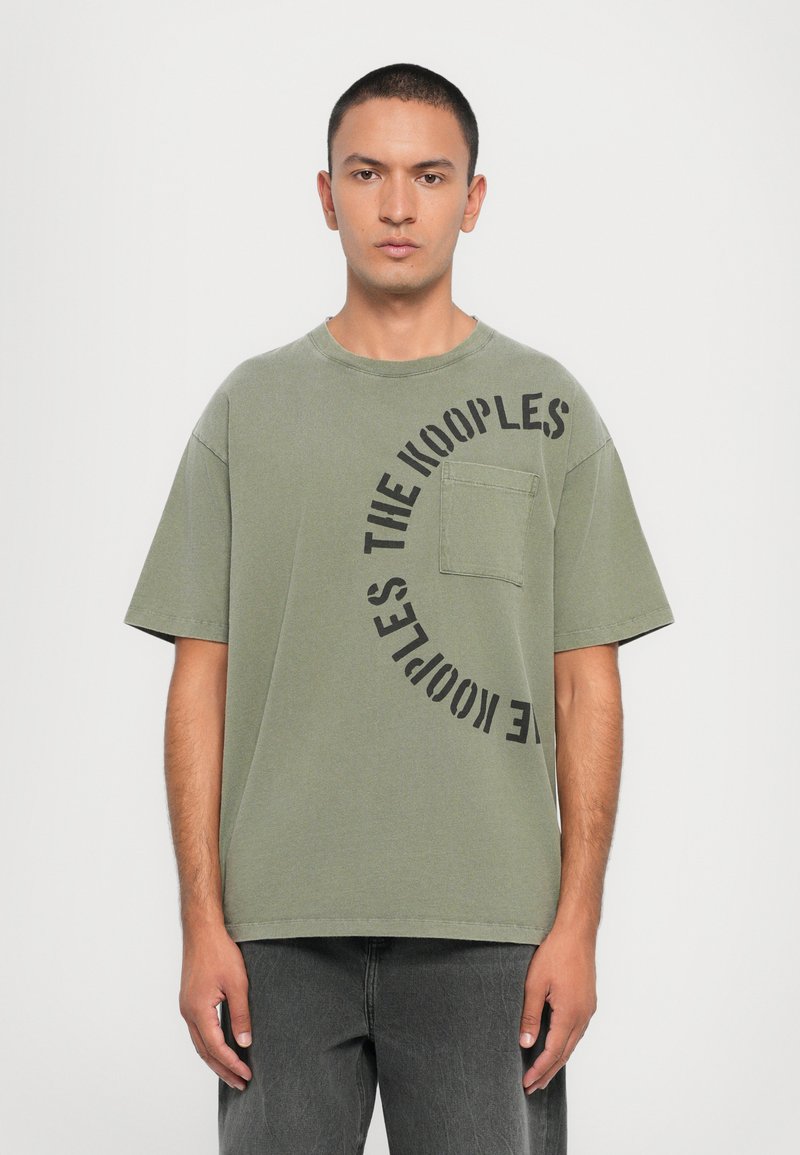 T-shirt oversized verde-azeitona com um bolso frontal, apresentando uma impressão gráfica de texto circular preto "THE KOOPLES" no lado esquerdo. Material em algodão.