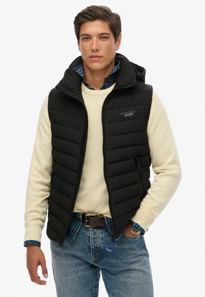 Young man wearing a black quilted vest over a cream sweater and blue plaid shirt with jeans, standing with one hand in his pocket.