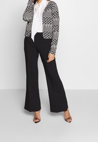 Black and white zigzag patterned cardigan, cropped design, paired with a white blouse and black wide-leg trousers. Black strappy heels included.