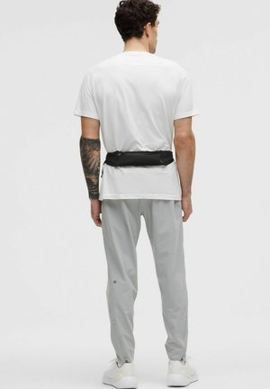 Outfit maschile composto da una camicia bianca a maniche corte, pantaloni grigi e una borsa a vita nera. Il tessuto sembra leggero e dal tocco morbido.