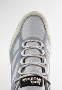 Szary but sportowy z teksturowaną cholewką, wyściełanym językiem, odblaskowymi detalami i szarymi sznurowadłami. Logo widoczne na języku.