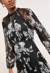 Rochie neagră cu imprimeu floral, mâneci lungi transparente, accente din dantelă și ținte argintii la talie. Prezintă modele mari de flori albe și gri.