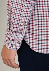 Camicia a quadri con un motivo rosso, blu navy e bianco; polsini con bottoni; tessuto leggero; orlo leggermente curvato. Outfit abbinato a pantaloni scuri.