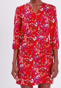 Robe fleurie rouge avec un col en V, des manches trois-quarts, et des motifs vibrants de fleurs roses et blanches avec des touches de vert. Tissu lisse.