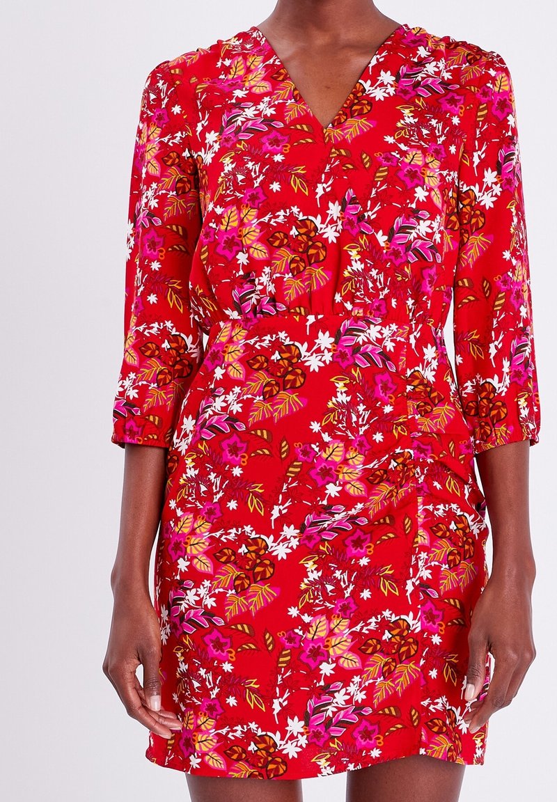 Robe fleurie rouge avec un col en V, des manches trois-quarts, et des motifs vibrants de fleurs roses et blanches avec des touches de vert. Tissu lisse.