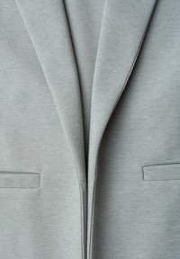 Close-up of a light gray blazer front showing lapels and two horizontal slit pockets on textured fabric.