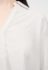 Γυναίκα που φοράει ανοιχτό μπεζ πουκάμισο με γιακά και μικρό ανάγλυφο λογότυπο τετράφυλλου λουλουδιού στο αριστερό στήθος.