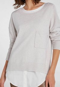 Hellgrauer gestrickter Pullover mit Rundhalsausschnitt, überschnittenen Schultern und einer Fronttasche, über einem weißen Hemd mit Kragen getragen.