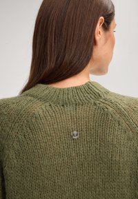 Maglione verde lavorato a maglia con una trama testurizzata, caratterizzato da un collo rotondo e un piccolo logo argentato sul retro. Il tessuto appare morbido e voluminoso.
