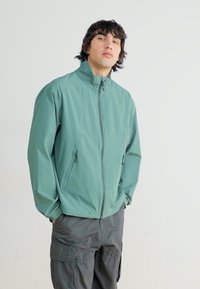 Green lightweight jacket with high collar, zip closure, and side pockets. Worn with dark cargo pants, featuring a smooth texture and modern design.