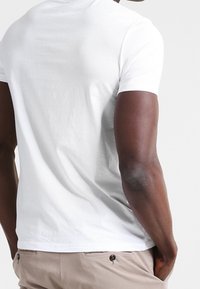 Uomo che indossa una semplice maglietta bianca a maniche corte e pantaloni beige, visto da dietro con le mani nelle tasche.