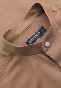 Chemise en tissu brun texturé avec un col montant et un bouton blanc unique. L'étiquette indique "ambre" dans un design noir et vert.