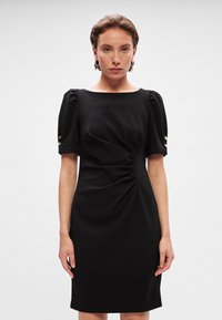 Crna uska haljina s kratkim puf rukavima, kvadratnim dekolteom, bočnim nabranim detaljima i zlatnim dugmadima na rukavima. Glatka tekstura tkanine.