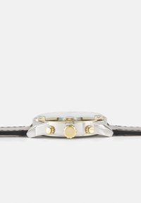 Ezüst polírozott tokú, három aranyszínű gombbal és fekete varrott bőrszíjjal rendelkező karóra oldalnézete fehér háttéren.