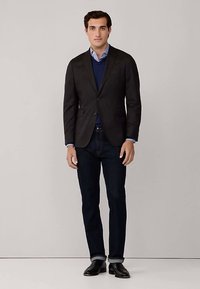 Blazer scuro, maglione blu, camicia azzurra, jeans scuri e scarpe nere indossati da una persona in piedi davanti a uno sfondo neutro.