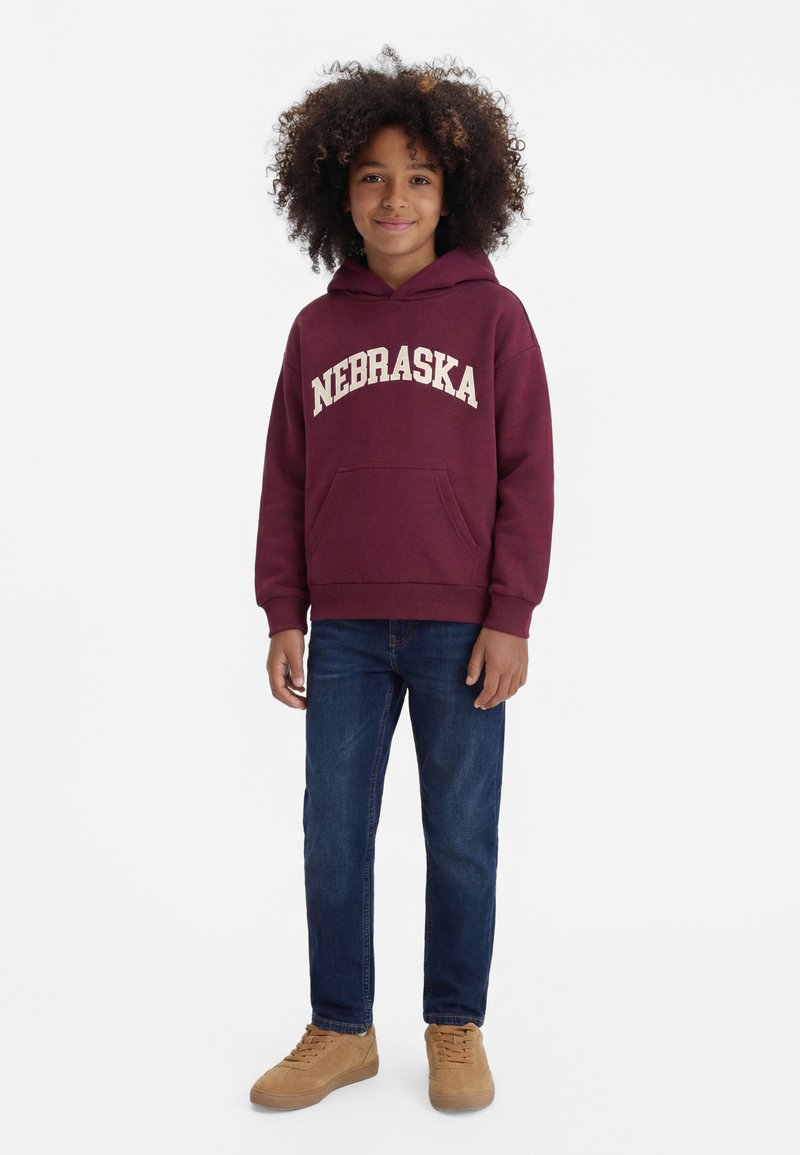 Kind mit lockigem Haar, das einen dunkelroten "Nebraska" Hoodie, blaue Jeans und beige Sneaker trägt, steht vor einem weißen Hintergrund.