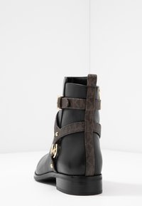 Botte à cheville en cuir noir avec une sangle à motif marron et des accents dorés. Présente un bout arrondi et un design à talon plat.