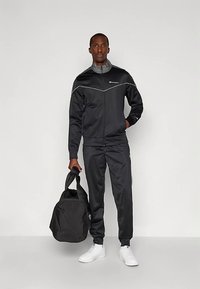 Melns sporta tērps ar pelēkām detaļām, rāvējslēdzēja jaka un bikses. Modelis tur melnu sporta somu, valkā baltas sporta kurpes. Vienkāršs, sportisks dizains.