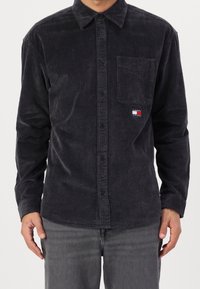 Camicia a bottoni in velluto a coste nera con taschino sul petto, caratterizzata da sottili righe verticali e una piccola patch con logo nei colori rosso, bianco e blu.