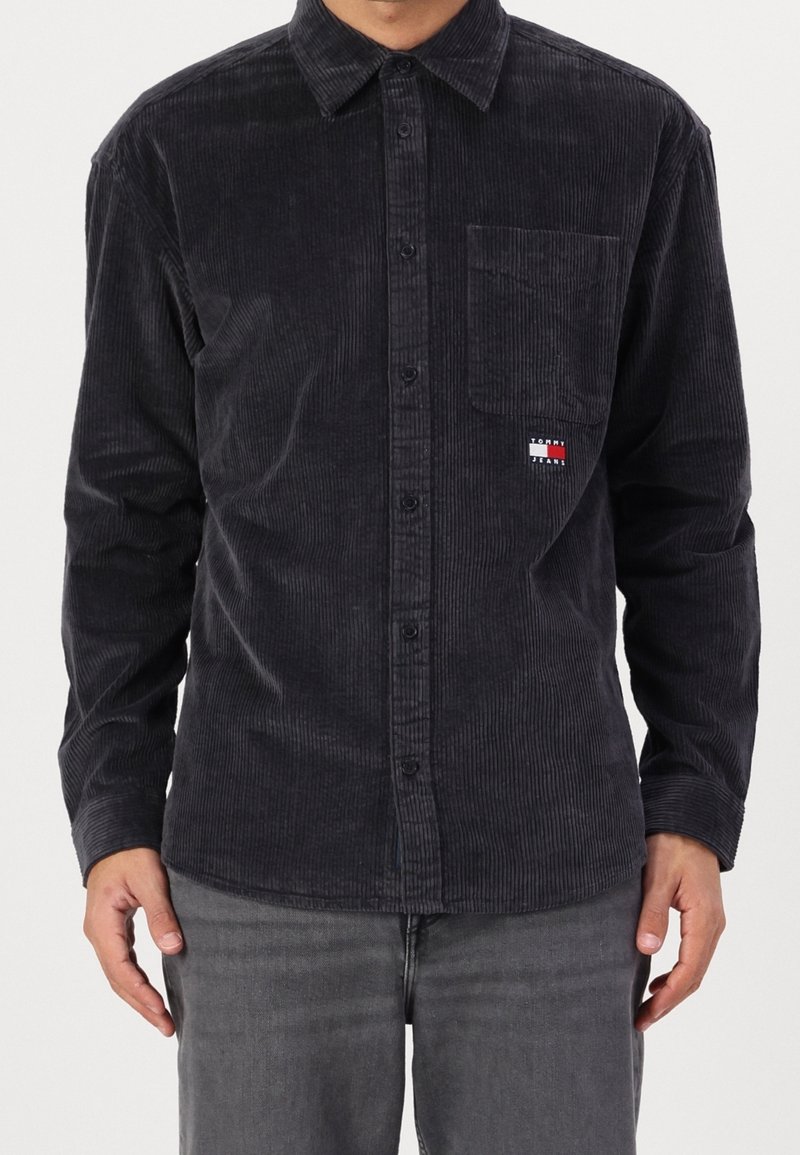 Camicia a bottoni in velluto a coste nera con taschino sul petto, caratterizzata da sottili righe verticali e una piccola patch con logo nei colori rosso, bianco e blu.