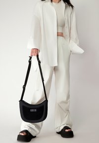 Borsa nera a spalla con una forma curva, tessuto texturizzato e tracolla regolabile, tenuta con una mano. Modella che indossa una camicia oversize bianca e pantaloni.