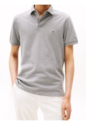 Hombre con una camisa polo gris claro de manga corta con un pequeño logo en el pecho y pantalones blancos, manos en los bolsillos.
