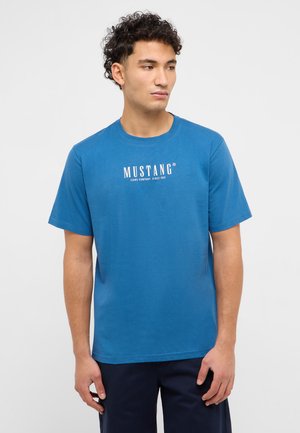 Mann mit lockigem Haar, der ein blaues Mustang-T-Shirt und dunkle Hosen trägt, steht vor einem einfarbigen hellen Hintergrund und schaut nach rechts.