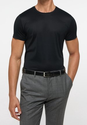 Mann trägt ein schwarzes, eng anliegendes T-Shirt mit kurzen Ärmeln, gesteckt in graue Anzugshosen mit schwarzem Gürtel, steht mit einer Hand in der Tasche.
