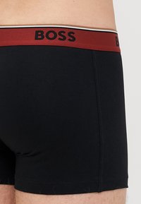 Boxeri scurți negri din bumbac cu un elastic roșu. Elasticul prezintă numele mărcii "BOSS" în litere negre și un accent cu dungi.