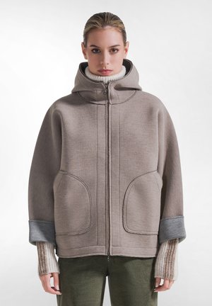 Mujer que lleva una chaqueta con capucha y cremallera color topo, con grandes bolsillos delanteros y mangas remangadas sobre guantes de punto sin dedos y pantalones verdes.